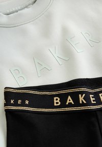 Svetlo siva kapuca s povišenim tekstom "BAKER" i crnim pasom s zlatnim natpisom "BAKER" i dekorativnim prugama.