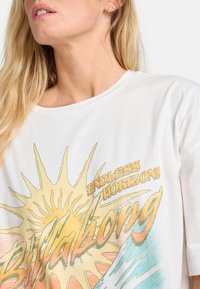 White t-shirt with a boat graphic, featuring a yellow sun and blue waves. Soft fabric with a relaxed fit and round neckline.