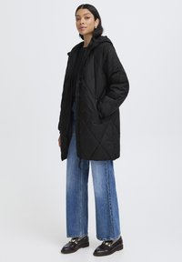 Veste longue noire matelassée avec une capuche, dotée d'une fermeture éclair et de poches latérales, portée sur un haut noir et un jean large bleu clair.