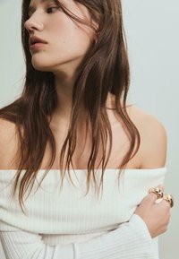 Biała bluzka z dekoltem off-the-shoulder, prążkowana, z długimi rękawami. Modelka z długimi brązowymi włosami prezentuje złote pierścionki na prawej ręce na jasnym tle.