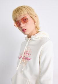 Balta gobtuvė su rožiniu "Nike Sportswear" grafiku, raišteliais ir priekinėmis kišenėmis; derinama su stačiakampio formos rožiniais saulės akiniais.