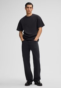 Schwarzes Baumwoll-T-Shirt mit kurzen Ärmeln, lockere Passform, kombiniert mit dunklen Jeans und schwarzen Schuhen, stehende Figur mit Händen in den Taschen.