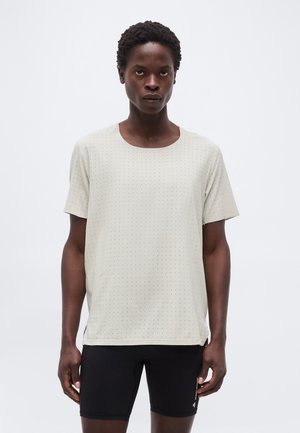 Άνδρας που φοράει μπεζ κοντομάνικο πουκάμισο με μικρό πουά μοτίβο και μαύρο σορτς ποδηλασίας σε απλό λευκό φόντο.