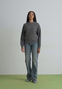Grå bomullströja med rund halsringning och ribbad nederkant, kombinerad med ljusblå utsvängda jeans. Modellen står i en neutral miljö.