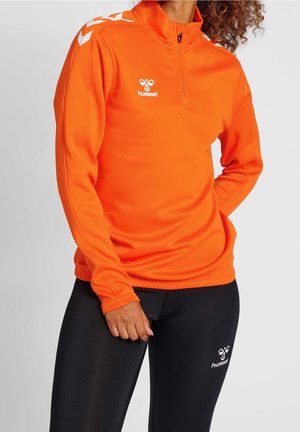 Persona que lleva una camiseta naranja de manga larga con cremallera y leggings negros, ambos con logos blancos de Hummel y franjas de cheurón en las mangas.