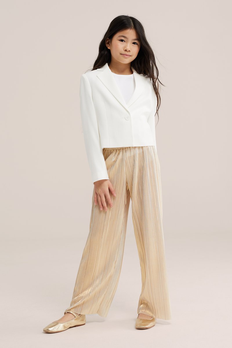 Niña joven con pelo largo y oscuro, que lleva una blazer blanca, top blanco, pantalones anchos plisados dorados y bailarinas doradas, de pie sobre un fondo neutro.