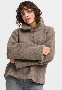 Brun fleecepullover med høj krave, kvart lynlås, brede ærmer og struktureret stof. Afsluttet med en buet kant.