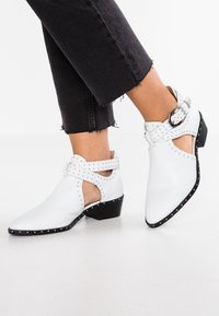 Botas de tobillo de cuero blanco con un diseño recortado, tachuelas plateadas en los bordes y un tacón bajo tipo bloque. Incluye un cierre de hebilla lateral.