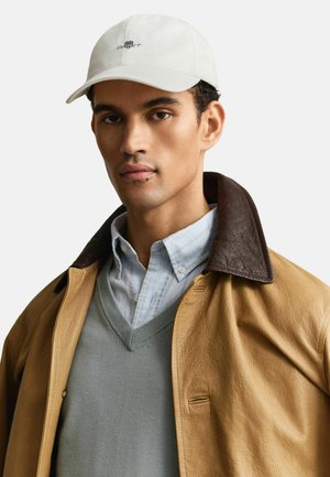 Mann mit weißer Mütze, hellblau gestreiftem Hemd mit Knöpfen, grauem V-Ausschnittpullover und beige Lederjacke mit dunkelbraunem Kragen.