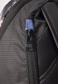 Esquina de una mochila negra que presenta un material texturizado, un panel de malla y una cremallera con un tirador de metal negro etiquetado "ROXY."