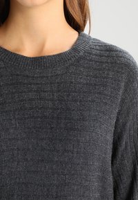 Šedý pletený svetr s kulatým výstřihem, který má texture a žebrovaný design. Základní materiál vypadá měkký a teplý.
