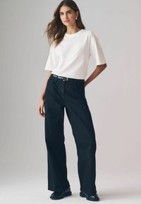 Weißes kurzärmliges T-Shirt kombiniert mit hoch taillierten, weit geschnittenen schwarzen Jeans, dazu ein schwarzer Gürtel mit einer goldenen Schnalle und schwarze Schuhe.