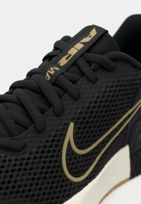Crna atletska cipela s strukturiranim mrežastim gornjištem, zlatnim Nike logom, ravnim crnim vezicama te bijelim i gumiranim potplatom.