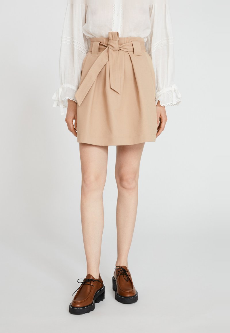 Beiger A-Linien-Rock mit einem geknoteten Gürtel, kombiniert mit braunen Schnürschuhen und einer hellen Bluse mit Rüschensleeves. Glatte Textur.