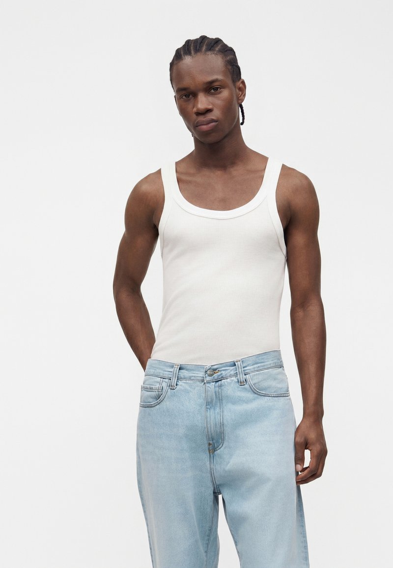 Young man with braided hair wearing a white sleeveless ribbed tank top and light blue jeans standing against a plain background.