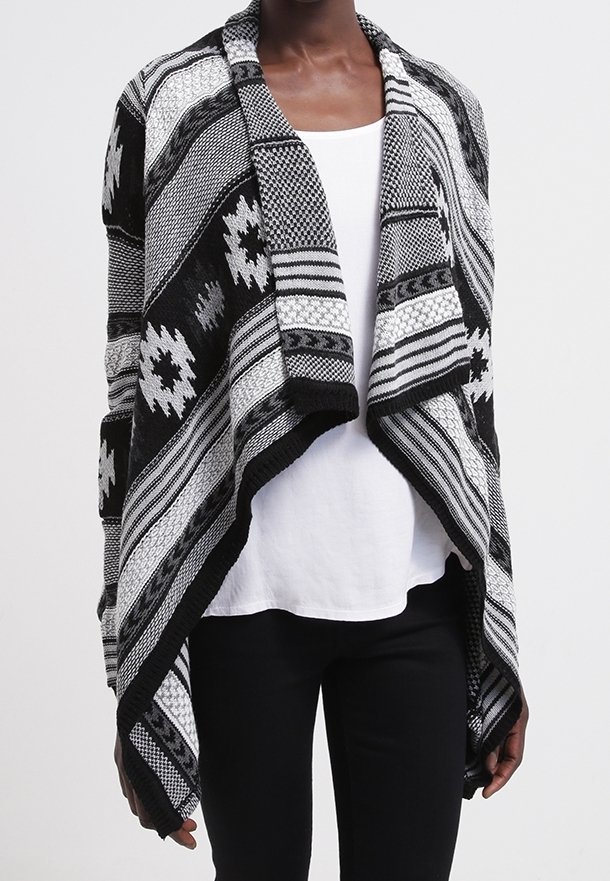 Cardigan de punto abierto con estampado en blanco y negro, usado sobre una camiseta blanca y pantalones negros por una persona de pie.