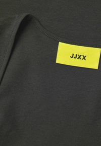 Melns audums ar gludu tekstūru un redzamu apkakli; uz tā ir spilgti dzeltens etiķete ar uzrakstu "JJXX" melnā krāsā.