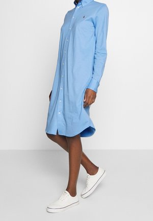 Femme portant une robe-chemise bleu clair à manches longues boutonnée, avec des baskets blanches, debout devant un fond blanc uni.