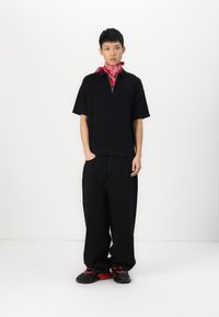 Black short-sleeve shirt with a zip neck, paired with loose black trousers. A red bandana is worn around the neck. Black sneakers with red accents.