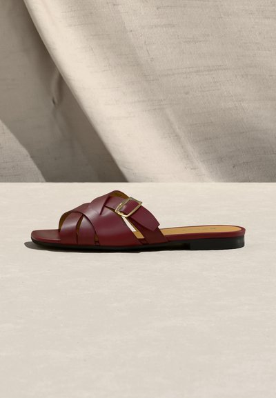 Sandale plate en cuir bordeaux avec larges brides tissées et une boucle dorée, posée sur une surface beige clair avec un fond de tissu drapé.