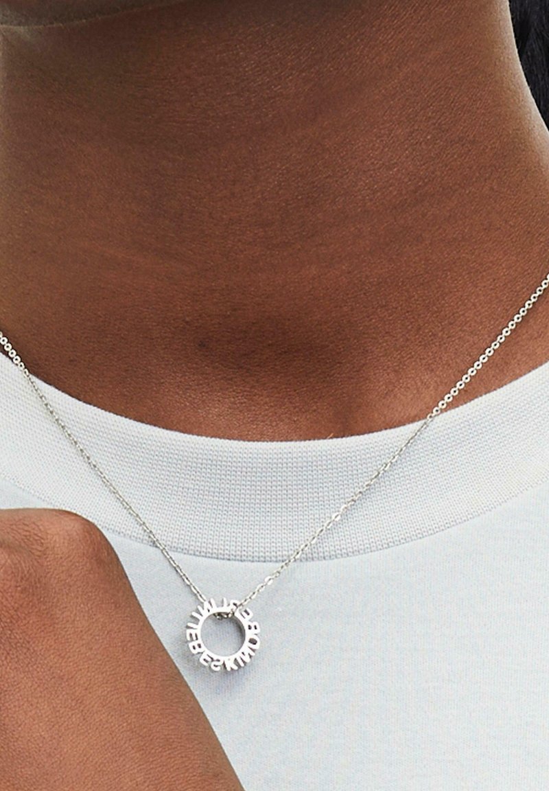 Collier en argent avec un pendentif circulaire arborant des lettres en relief. La chaîne est fine avec une surface texturée, portée sur un vêtement bleu clair.