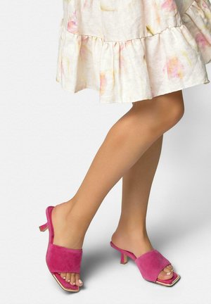 Piernas con una falda ligera de flores y mules de tacón abiertos color rosa brillante con punta cuadrada sobre un fondo blanco.