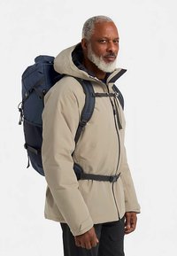 Giacca impermeabile beige con cappuccio, cerniere e vita aderente. Zaino blu scuro con dettagli in rete e spallacci regolabili.