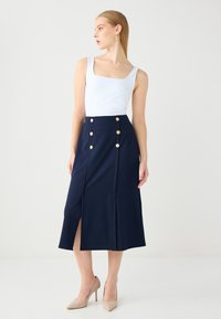 Gonna midi blu navy con spacco frontale, decorata con bottoni dorati e vita alta. Abbinata a un top senza maniche bianco e décolleté nude a punta.