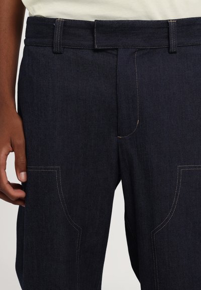 Dunkelblaue Denim-Hosen mit geradem Schnitt, versehen mit sichtbaren Nähten, Gürtelschlaufen und Fronttaschen. Die Textur wirkt robust und langlebig.