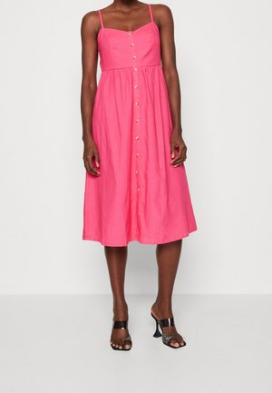 Frau, die ein ärmelloses pinkfarbenes Midi-Kleid mit vorderen Knöpfen und schwarzen Sandalen mit hohen Absätzen trägt, steht vor einem einfarbigen hellen Hintergrund.