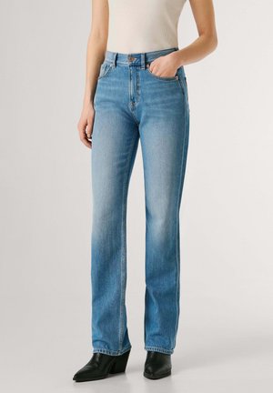 Donna che indossa jeans dritti a gamba larga azzurri con vita alta, abbinati a stivaletti neri con tacco e una canotta bianca senza maniche.