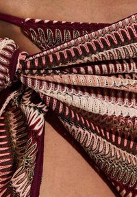 Primer plano de tela de ganchillo en tonos granate y beige, atada en un nudo sobre la piel, mostrando patrones tejidos y textura detallada.