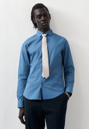 Άντρας με σκούρα dreadlocks, που φοράει μπλε πουκάμισο, ανοιχτόχρωμη γραβάτα και σκουρόχρωμο παντελόνι, στέκεται σε έναν λευκό τοίχο με το ένα χέρι στην τσέπη.