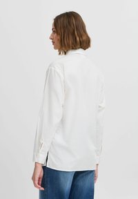 Chemise blanche à manches longues avec col, poignets boutonnés et fentes latérales, en tissu lisse. Portée avec un jean en denim bleu.