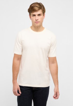 Junger Mann mit hellbraunem Haar, trägt ein schlichtes, cremefarbenes T-Shirt mit kurzen Ärmeln und dunkle Jeans, steht vor einem neutralen Hintergrund.