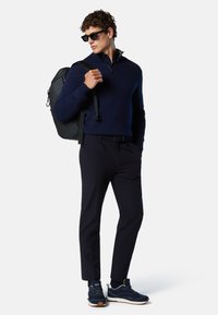 Pullover in maglina blu navy con collo a mezza zip, pantaloni neri e sneakers blu navy. Zaino nero sulla spalla sinistra, dettagli aggiuntivi comprendono occhiali da sole.