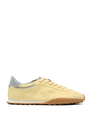 Sneaker gialla a profilo basso con tallone grigio chiaro, chiusura frontale con lacci e suola in gomma testurizzata rivolta a destra su sfondo bianco.