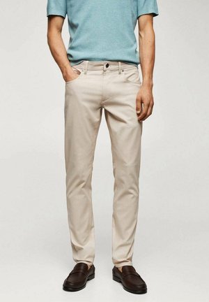 Mężczyzna w beżowych spodniach o dopasowanym kroju, jasnoniebieskim krótkim swetrze i ciemnobrązowych mokasynach, stojący z jedną ręką w kieszeni.