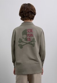 Tricou polo verde măsliniu cu mânecă lungă, cu o grafică pe spate reprezentând un craniu și oase încrucișate în verde închis, cu broderie roz.