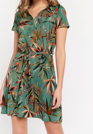 Grünes Kleid mit kurzen Ärmeln, orange-braun-schwarzem tropischem Blattmuster und passendem Stoffgürtel, der an der Taille gebunden ist.