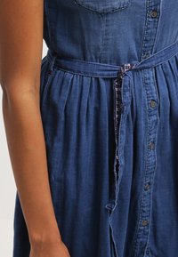 Sukienka dżinsowa z dopasowaną górą, plisowaną spódnicą i krótkimi rękawami. Posiada materiałowy pasek i zapięcie na guziki z przodu. Kolor ciemnoniebieski.
