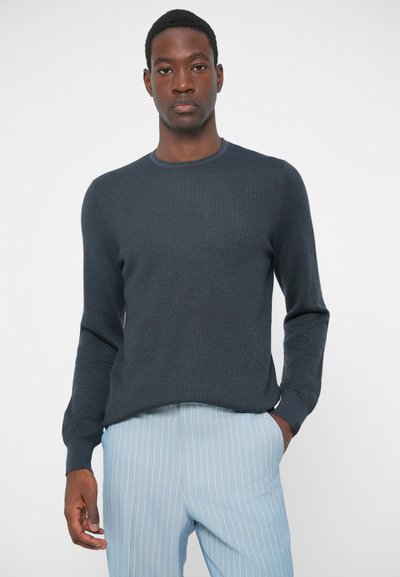 Dunkelblauer, gerippter Strickpullover mit Rundhalsausschnitt und langen Ärmeln, kombiniert mit hellblauen, gestreiften Hosen. Glatte Textur über die gesamte Oberfläche.