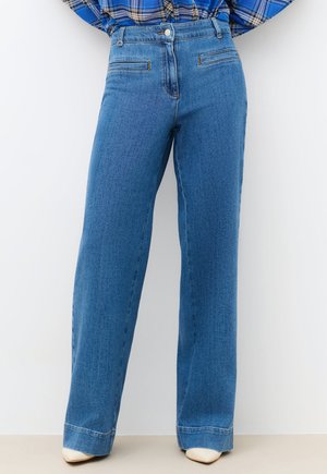 Frau trägt hoch taillierte gerade geschnittene blaue Jeans und weiße spitze Schuhe, kombiniert mit einem in die Hose gesteckten blau karierten Hemd.