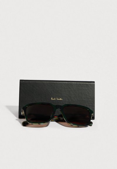 Γυαλιά ηλίου με πράσινο πλαίσιο από φλοίδα χελώνας και σκούρους φακούς, τοποθετημένα μπροστά σε μια μαύρη, υφασμάτινη θήκη με χρυσά γράμματα "Paul Smith".