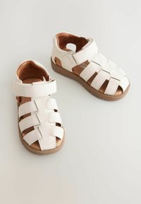 Hvide canvas sandaler med lukket tå-design, der har krydsspiraler og velcro-lukning. Tekstureret overflade med en brun gummisål.