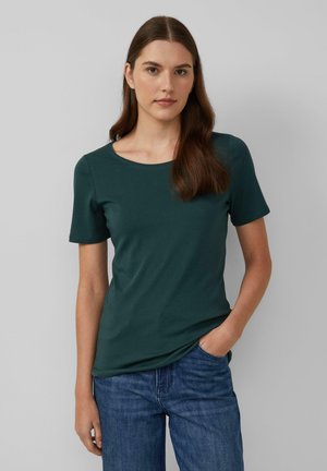 Jeune femme aux longs cheveux bruns portant un t-shirt à manches courtes vert foncé et un jeans bleu, se tenant avec une main dans sa poche.