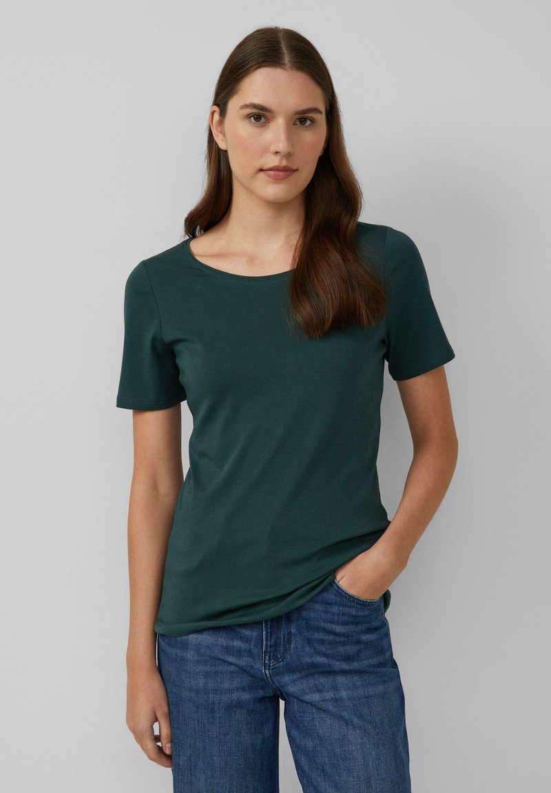 Jonge vrouw met lang bruin haar, in een donkergroene kortebloes met korte mouwen en blauwe jeans, staande met één hand in haar zak.
