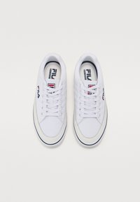 White sneakers with a canvas upper, flat laces, padded collar, and a rubber sole. Logo visible on the side and tongue in red and blue.
