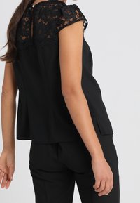 Svart topp med spetsdetaljer på ärmarna, har hög krage och en avslappnad passform. Bakstycket har en nyckelhålsdetalj och sidoflikar. Parats med svarta byxor.