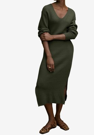 Femme portant une longue robe en maille côtelée vert olive avec un col en V et des ballerines à imprimé léopard, debout les bras croisés.
